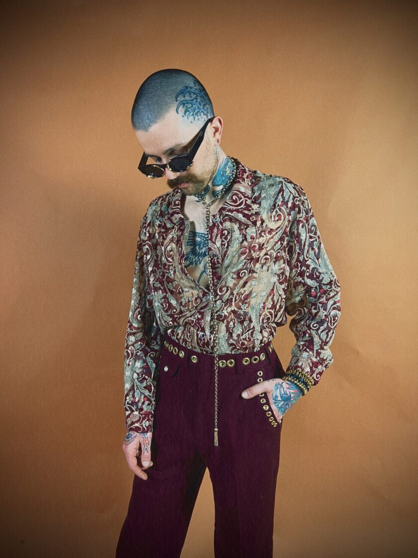 A person with tattoos and a shaved head, wearing sunglasses, a paisley shirt, and purple trousers, posing against a tan background.