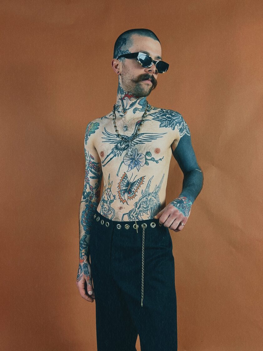 Man with extensive tattoos standing confidently against an orange background.