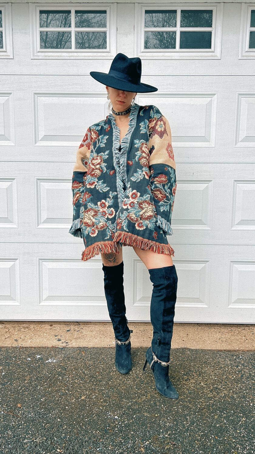 A person in a wide-brimmed hat, floral kimono, distressed jeans, and thigh-high boots standing in front of a garage door.