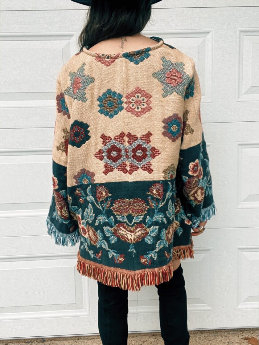 Woman standing with her back to the camera wearing a floral-patterned fringed poncho and black pants.