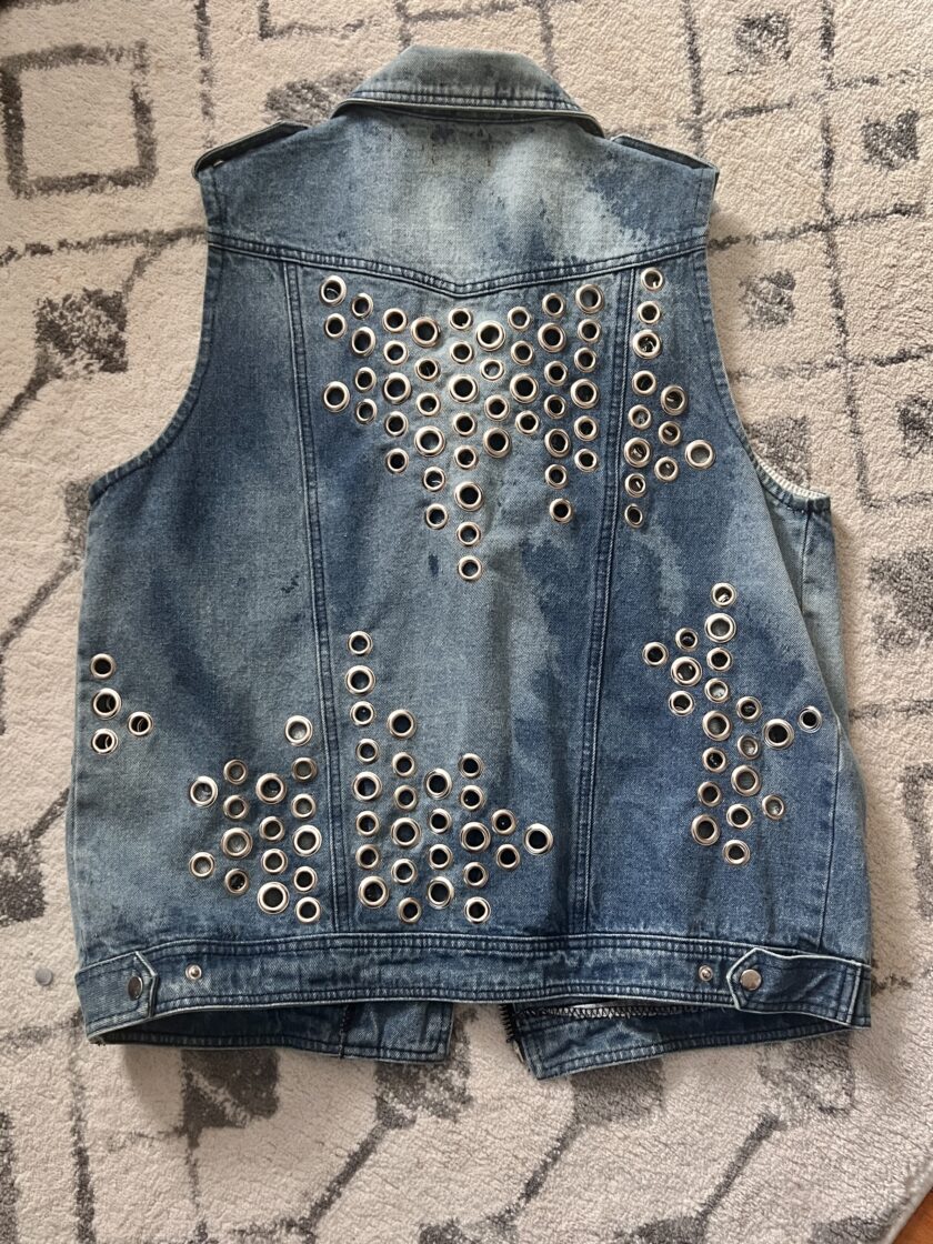 A denim vest with a pattern of metal eyelets laid out on a carpet.