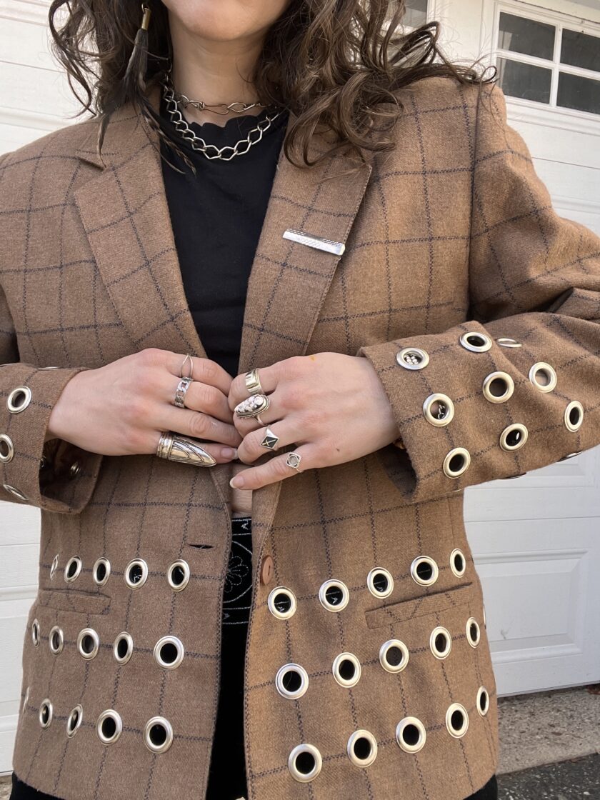A person wearing a plaid blazer adorned with numerous eyelets on the sleeves.