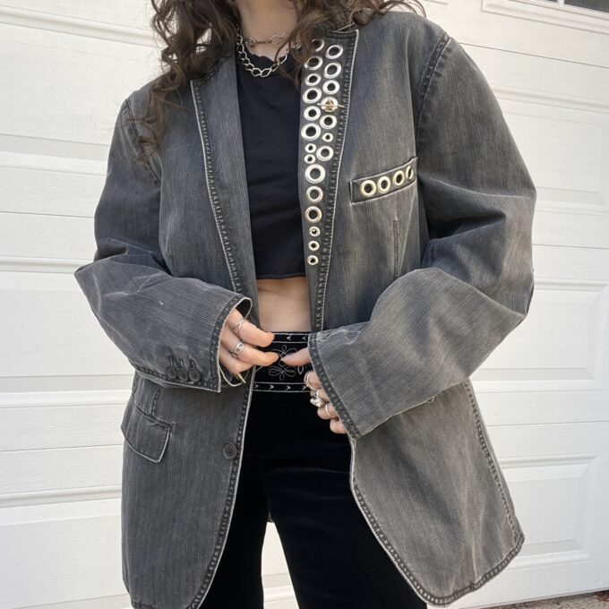 A person wearing a black crop top, black pants, and an oversized denim jacket with metal ring embellishments.