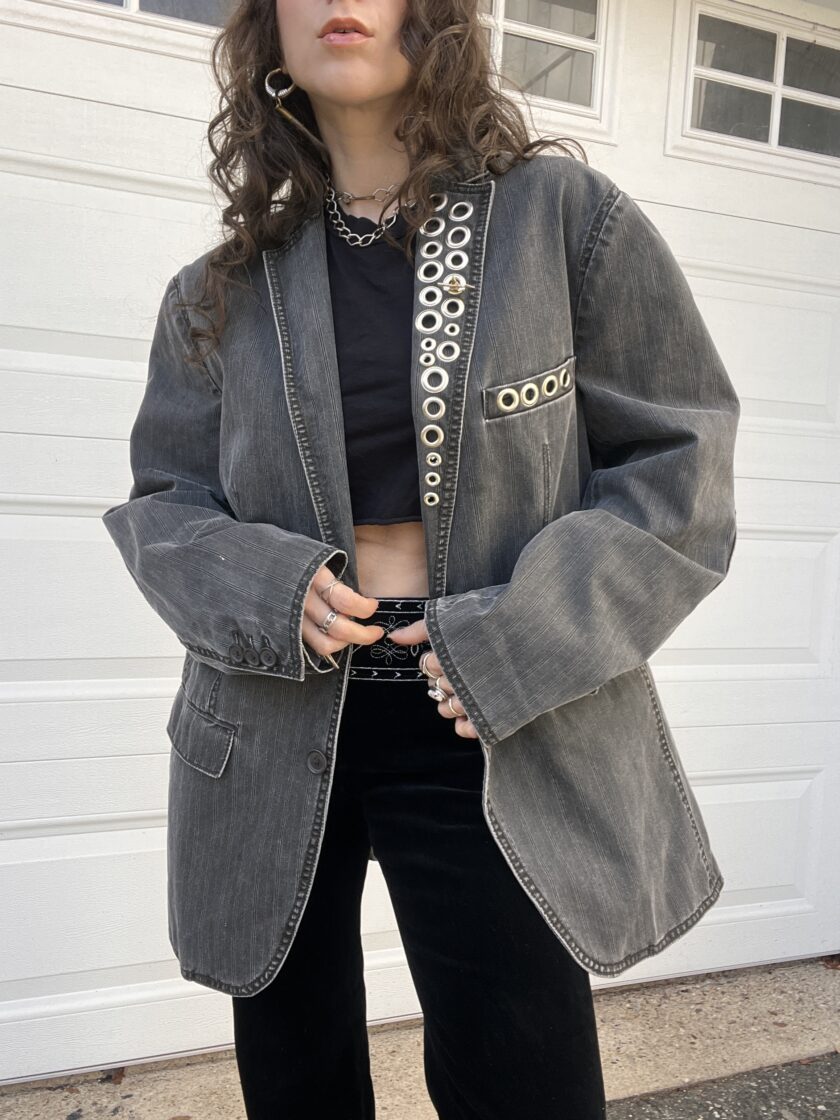 A person wearing a black crop top, black pants, and an oversized denim jacket with metal ring embellishments.