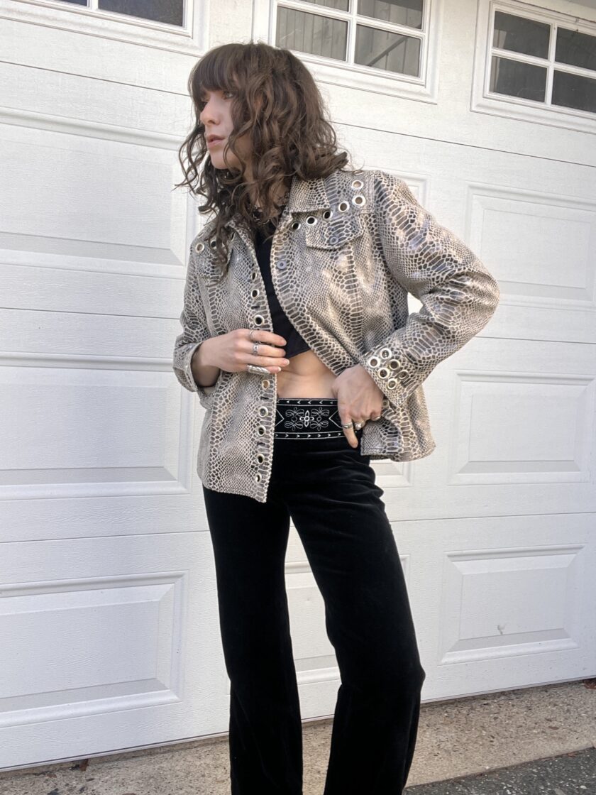 A person with curly hair wearing a metallic jacket and black pants standing in front of a white garage door.