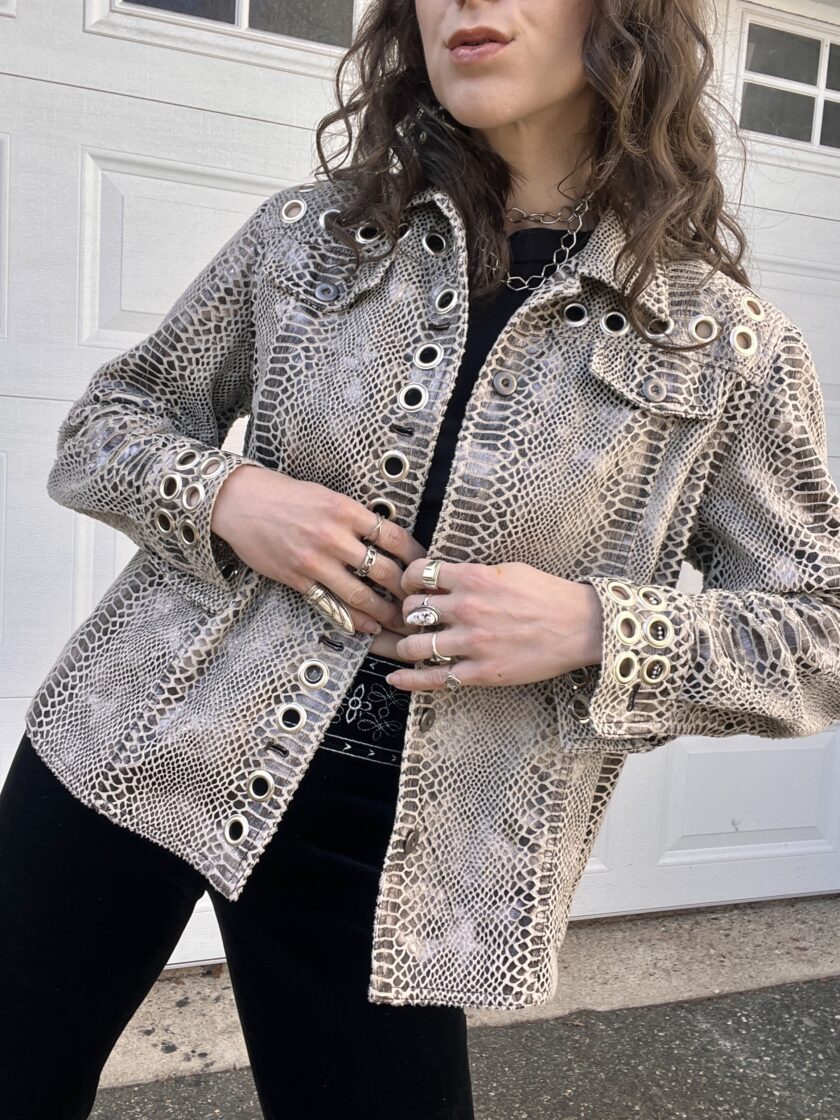 A person wearing a textured metallic jacket and black pants, accessorized with multiple rings and a chunky necklace.