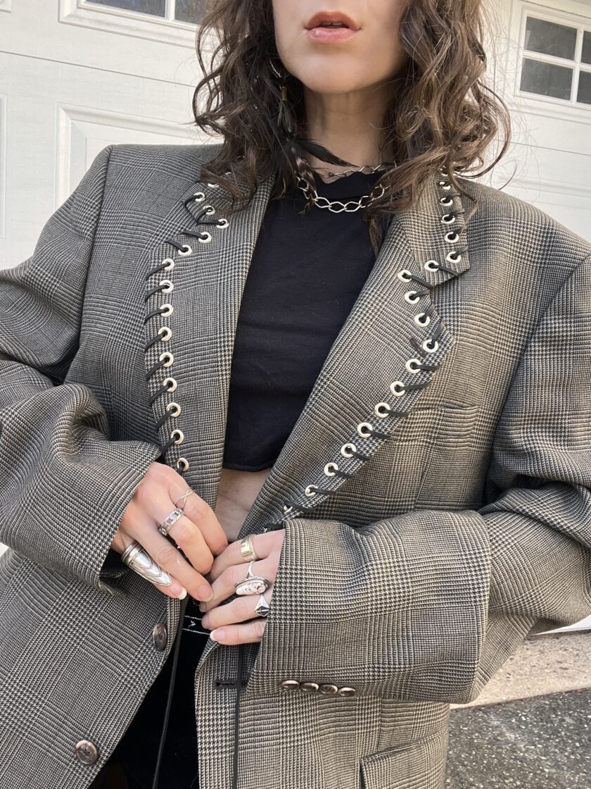 A person wearing a checkered blazer with metal eyelets and layered silver rings, accessorized with a chunky chain necklace.