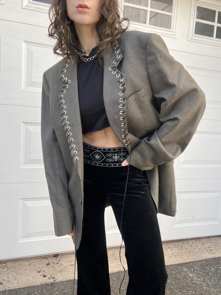 Person in a stylish outfit featuring a plaid oversized blazer with metal eyelet detailing, black crop top, high-waisted pants, and chunky necklace.