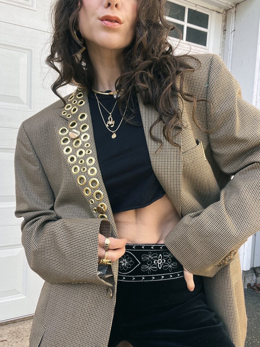 A person wearing a patterned blazer with embellished details, a black crop top, and a high-waisted skirt, accessorized with layered necklaces and rings.