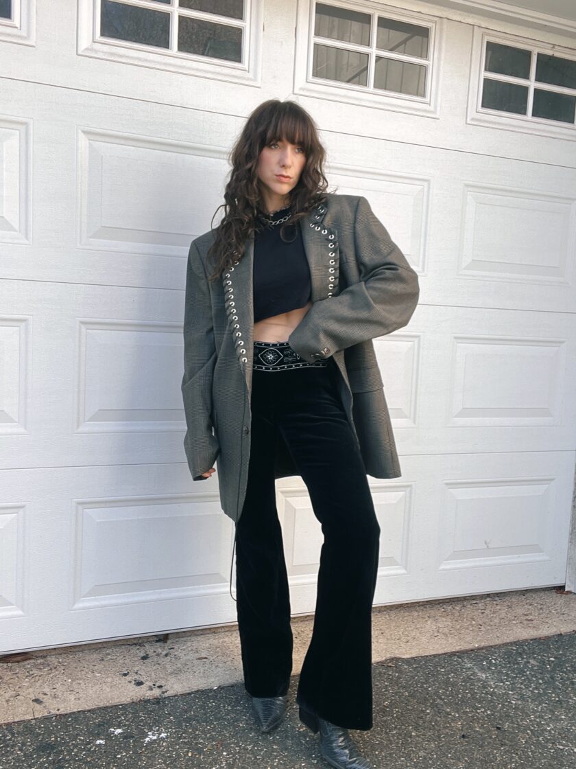 A person posing in front of a white garage door, wearing a black top and pants with a gray blazer and boots.