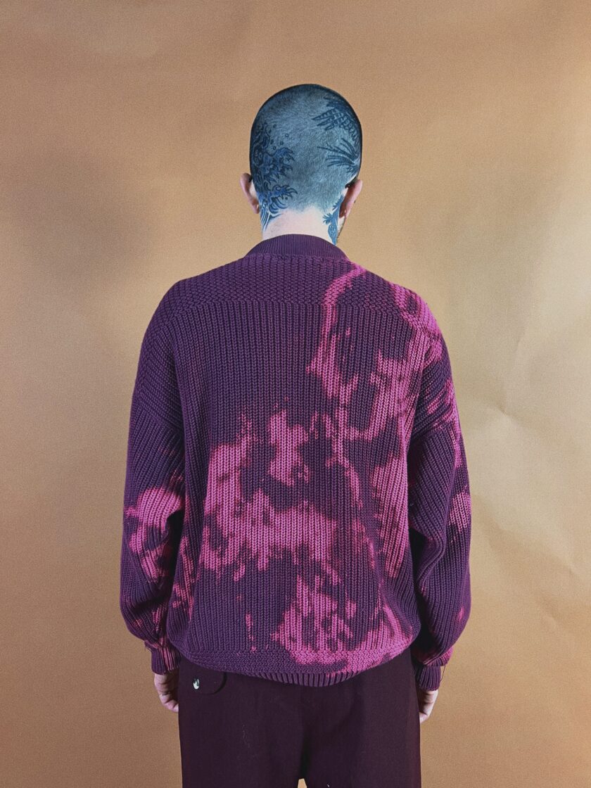 Individual with a tattooed head seen from behind, wearing a purple sweater with a pink pattern, standing against a tan backdrop.