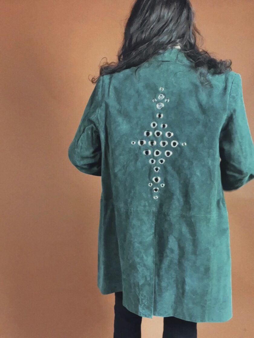 A person with long hair seen from behind wearing a teal suede jacket with a decorative pattern of metal eyelets on the back.