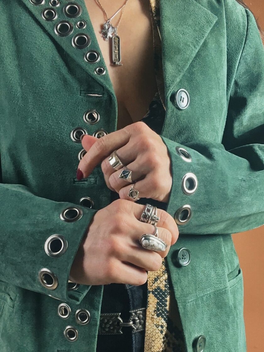 A person in a green jacket adorned with metal grommets, clutching the lapel with hands featuring multiple rings.