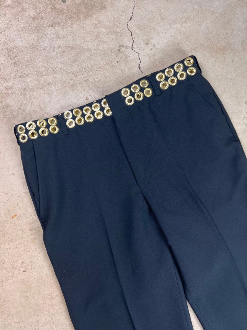 Navy trousers with an embellished waistband featuring circular gold-tone grommets on a concrete background.