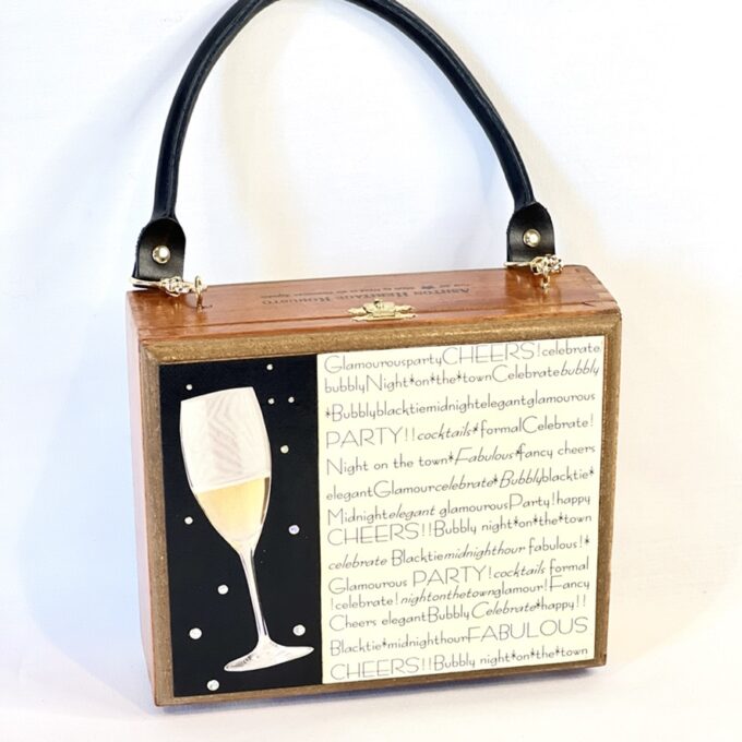 A square wood cigar box purse with a black handle with a Champagne glass image and text design, with celebratory words like "cheers!" and "party!".