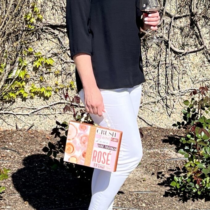 A person in white pants and a black top holding a box of rosé wine and a glass while standing outdoors.