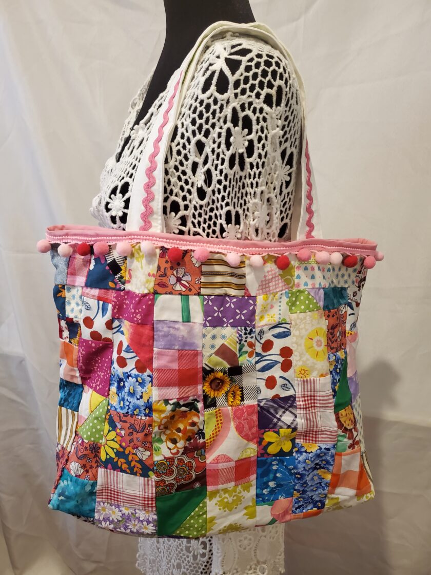 A colorful patchwork tote bag with a variety of floral and patterned fabrics, accented with a white lace overlay and pink trim, displayed on a mannequin.