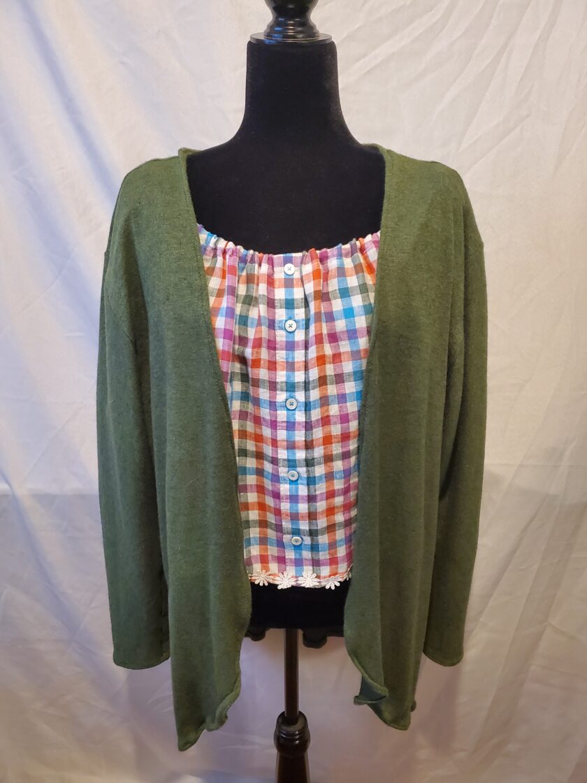 A mannequin displaying a multicolored checkered blouse with white buttons under a green cardigan against a neutral backdrop.