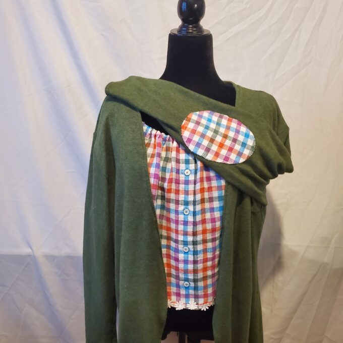 Mannequin dressed in a green cardigan over a plaid multicolored shirt, accented with a large oval brooch, against a white backdrop.