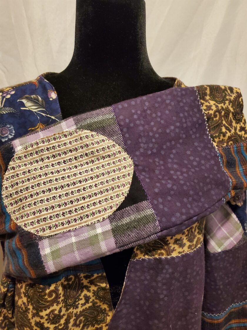 A mannequin displays a patchwork garment with a mix of floral, plaid, and solid fabric pieces in shades of purple, cream, and blue.