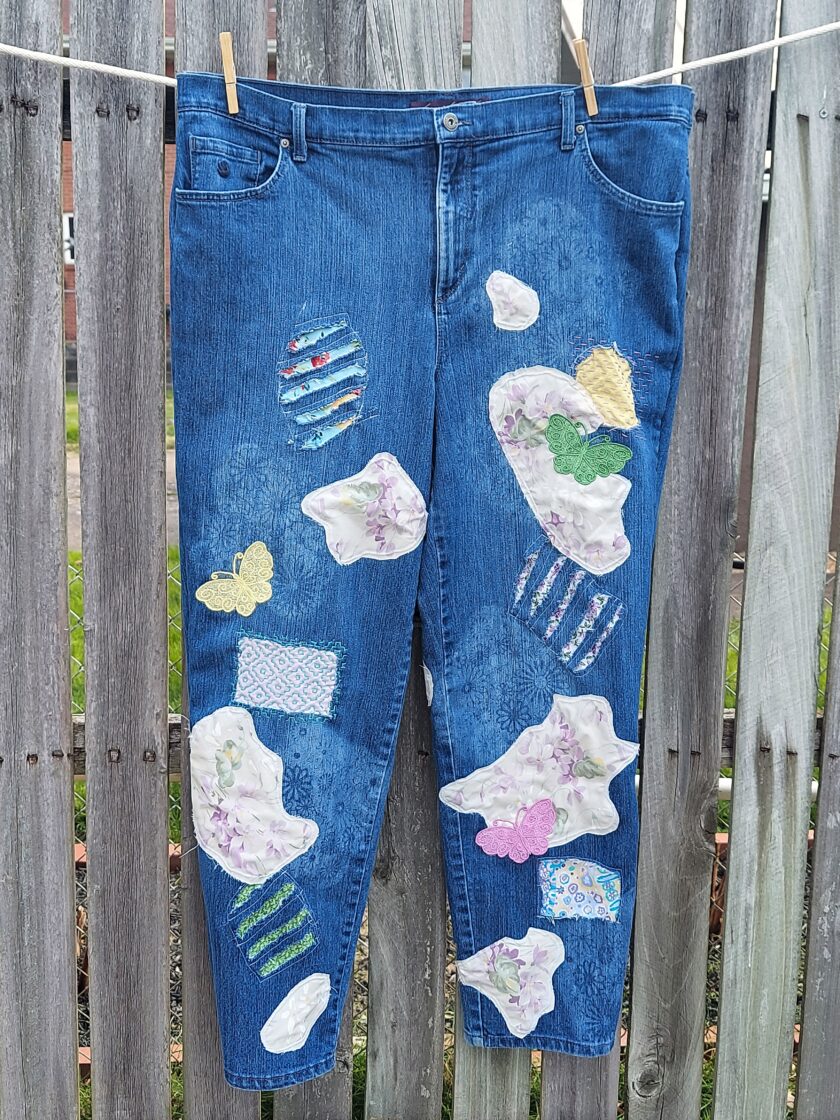 Blue jeans with floral patches hanging on a clothesline in front of a wooden fence.