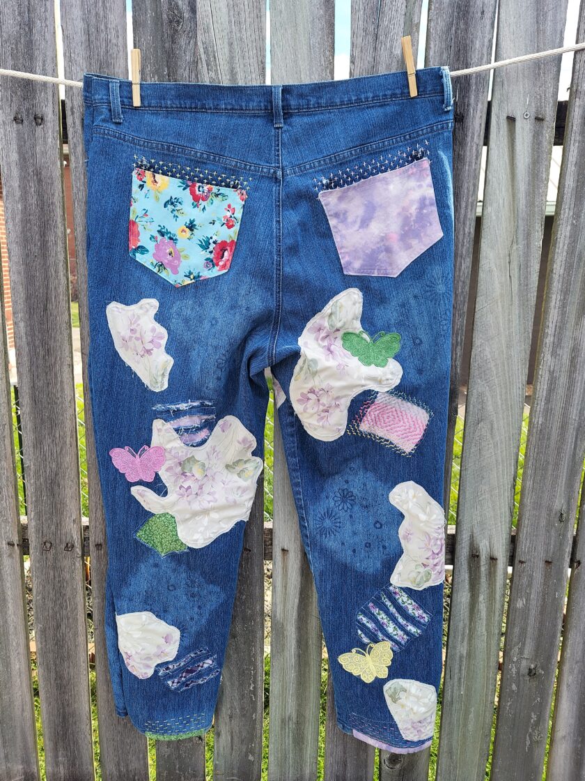 A pair of blue jeans with various floral and patterned fabric patches hanging on a clothesline outdoors.
