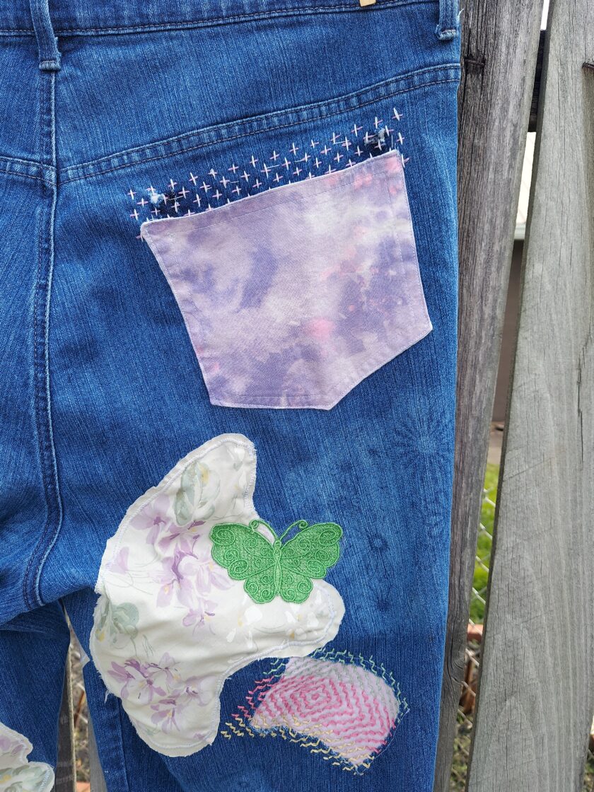 Close-up view of a pair of denim jeans with unique patches, including a floral pocket, embroidered details, and a butterfly embellishment.