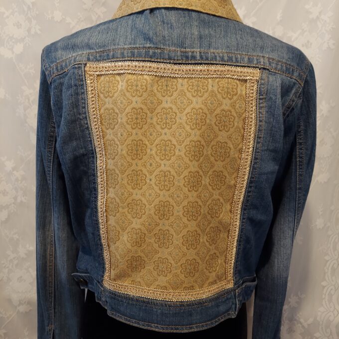 A denim jacket with a patterned beige fabric panel on the back and cuffs, displayed on a mannequin against a patterned background.