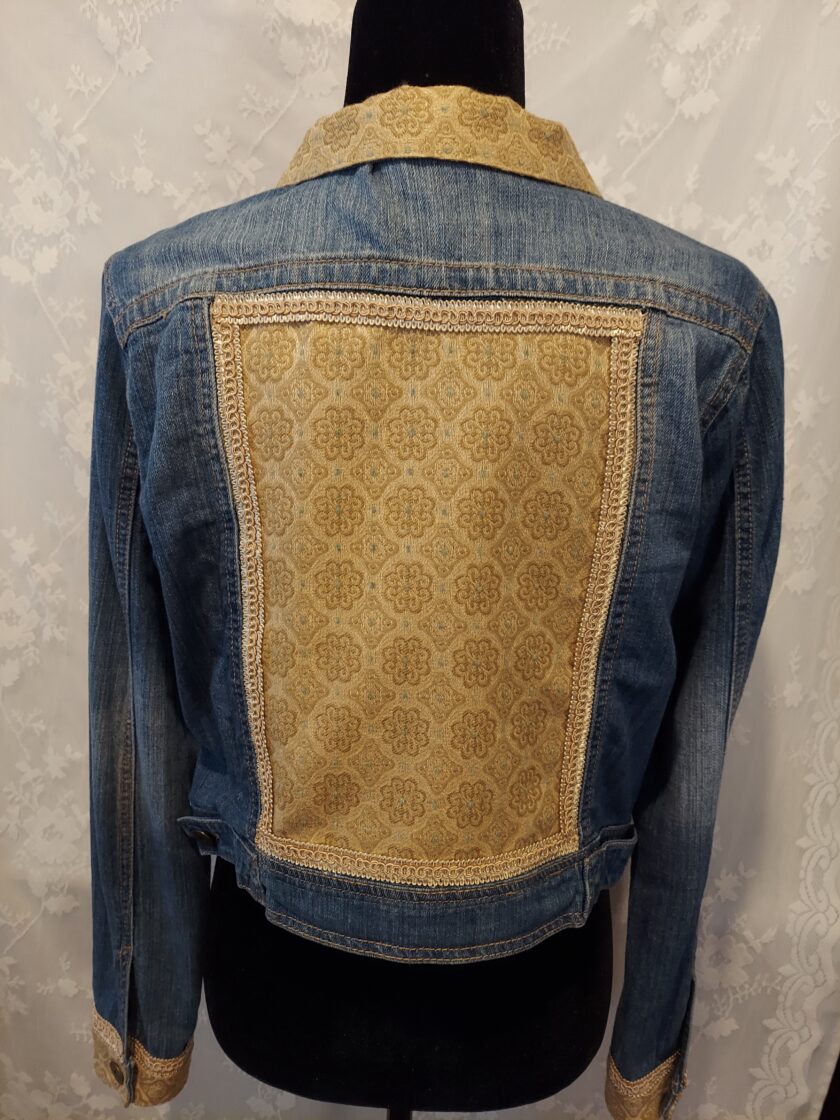 A denim jacket with a patterned beige fabric panel on the back and cuffs, displayed on a mannequin against a patterned background.