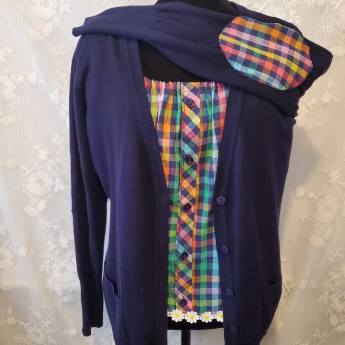 A mannequin displaying a quirky, patchwork cardigan with a plaid and floral pattern mix, draped over a dark, plain hoodie.