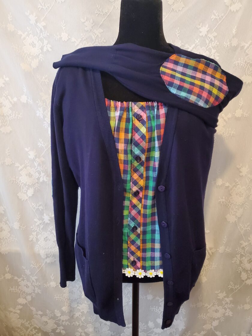 A mannequin displaying a quirky, patchwork cardigan with a plaid and floral pattern mix, draped over a dark, plain hoodie.