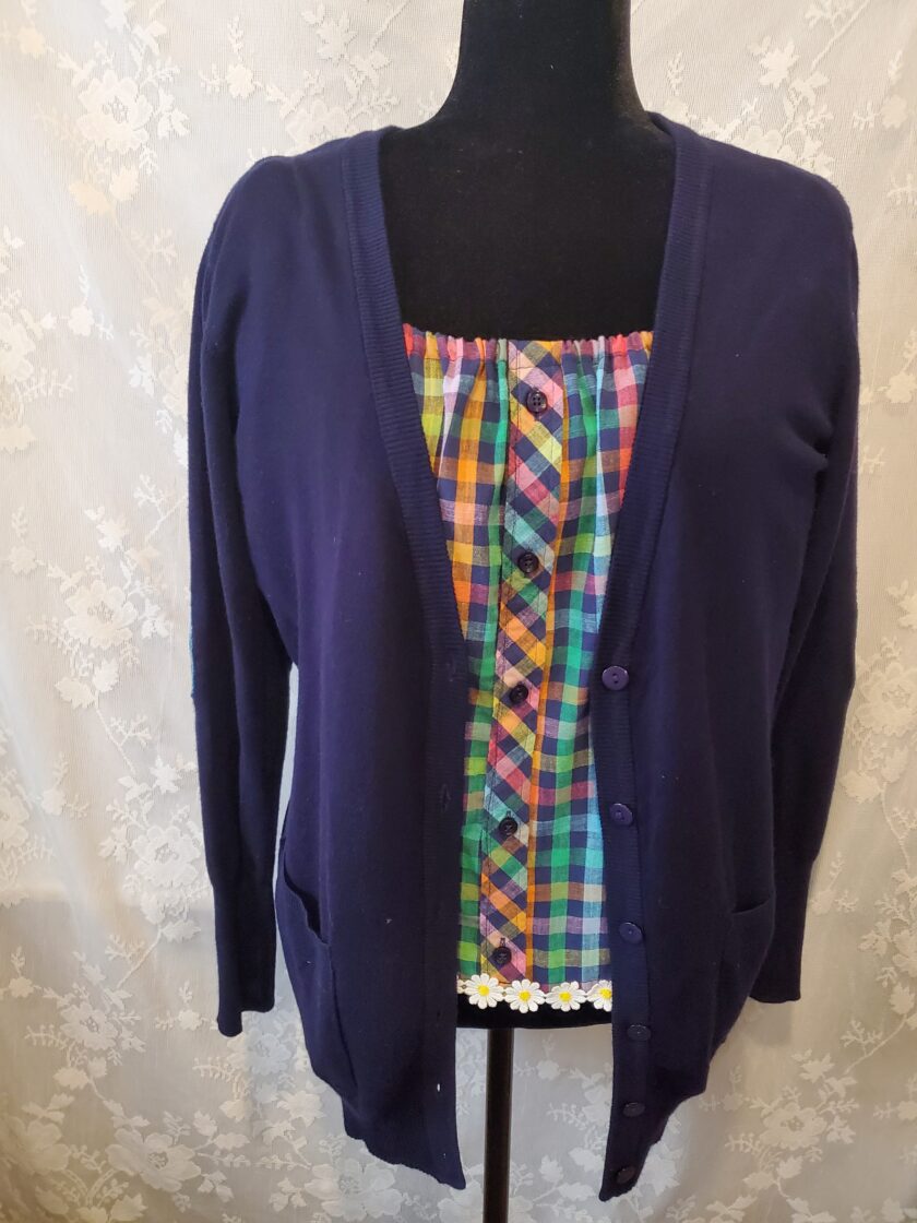 A navy blue cardigan with a multicolored plaid inset displayed on a mannequin against a patterned background.