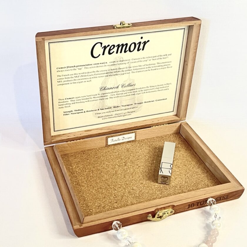 Open wooden box with a text label inside the lid, featuring a small metal clasp and a cork base, displayed with a pearl necklace and bracelet.