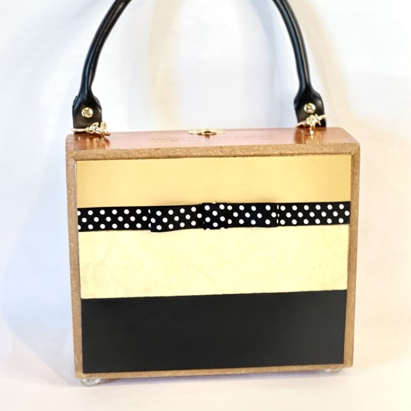 A square, box-style handbag with a striped design in ivory, black, and gold, featuring polka-dot ribbon and black handle