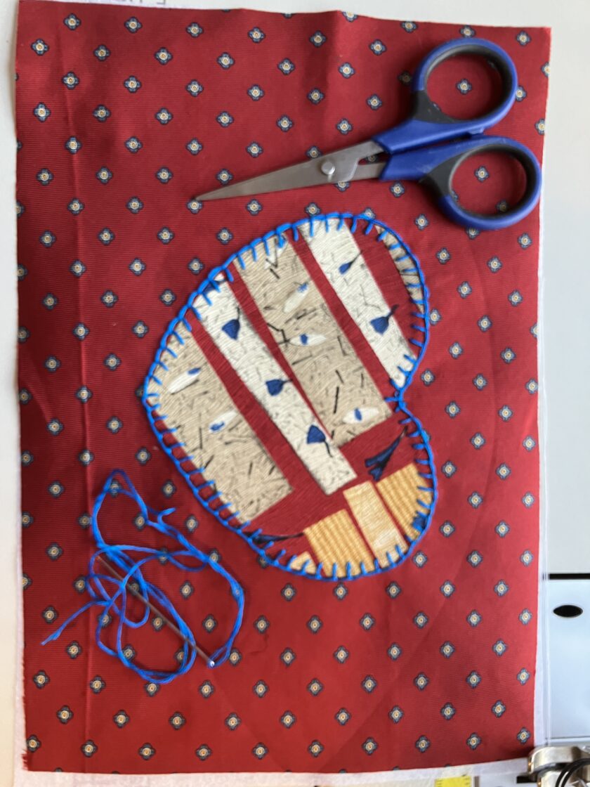 A craft project featuring a red fabric bookmark with a blue and white striped heart appliqué, alongside scissors and blue thread on a patterned background.