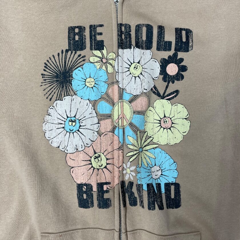 A beige hoodie featuring the phrase "be bold be kind" in black letters, decorated with colorful floral graphics and a peace symbol on the zipper line.