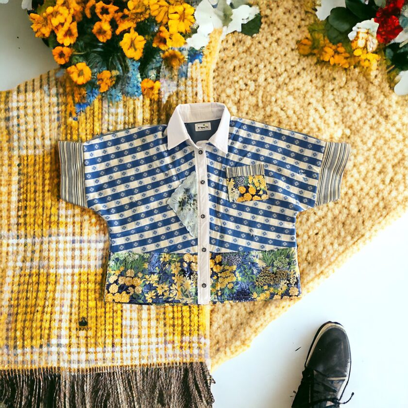 A patterned shirt laid flat on a yellow rug with colorful flowers and a black shoe at the bottom right corner.