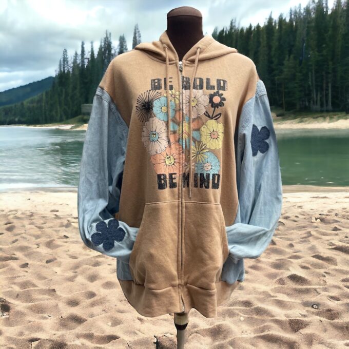 A hooded sweatshirt displayed on a mannequin at a sandy lakeside, featuring floral designs and the text "be bold be kind.