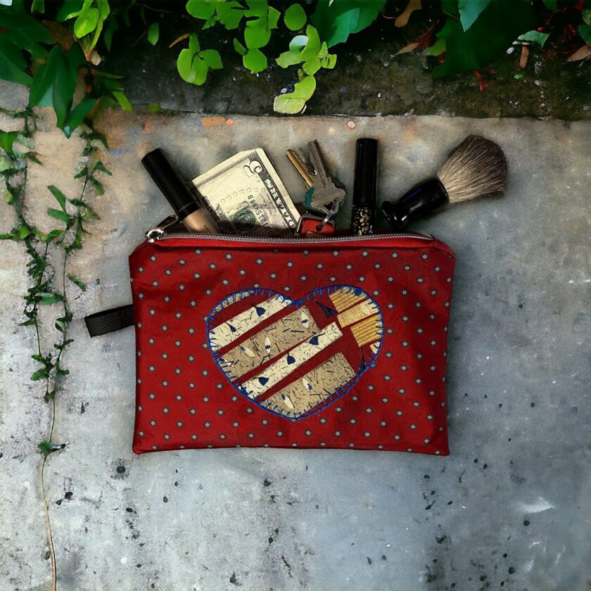 A red cosmetic bag with a heart design, unzipped to reveal cash, keys, and makeup items, rests on a stone surface surrounded by ivy.
