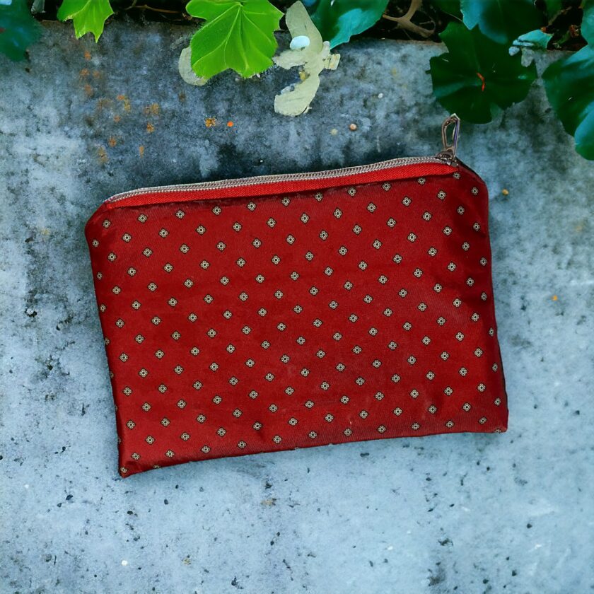 Red fabric pouch with a diamond pattern and a zipper, resting on a textured gray surface with green leaves and white flowers around it.