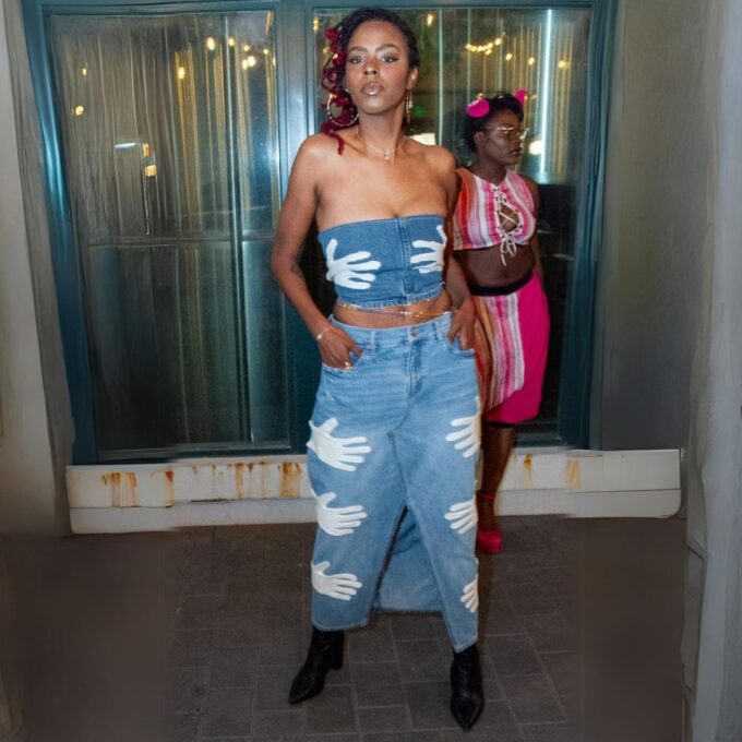 A woman in a denim outfit with handprint designs poses for a photo while another person is visible in the background.