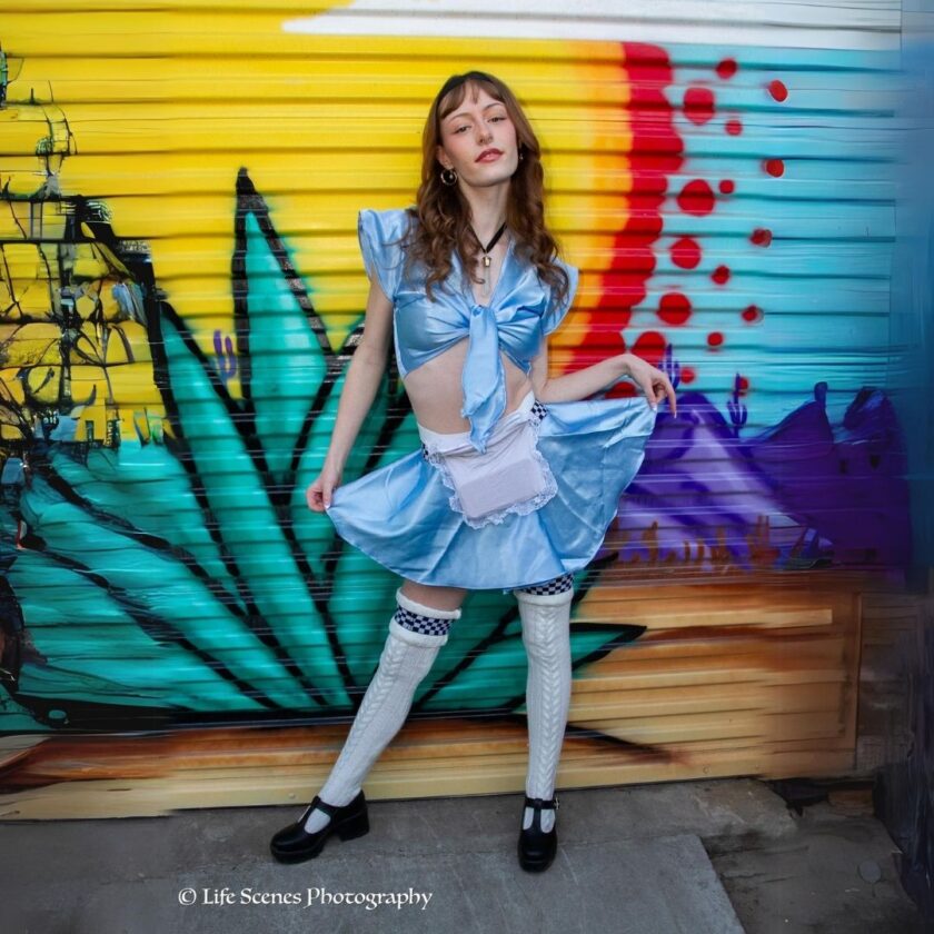 A person in an alice in wonderland costume posing against a colorful mural.