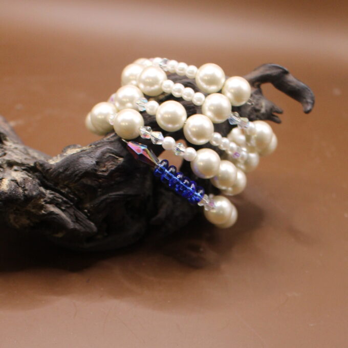 A pearl bracelet with a blue gemstone accent resting on a piece of driftwood against a neutral background.