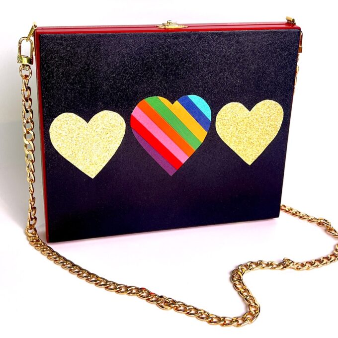 Black glitter covers a red box purse, with three hearts, in gold glitter and rainbow, with a gold chain strap.